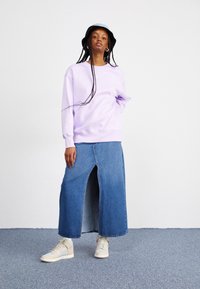 Sweatshirt lilás claro com decote redondo, combinado com uma saia maxi de denim com fenda alta e ténis bege. Chapéu de balde preto e tranças longas.