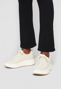 Creme platform sneakers med vævet tekstur, snørebåndsdesign og en tyk hvid sål. Accentueret med beige detaljer langs kanten.