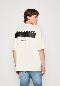 Kremfarget oversized T-skjorte med svart grafisk trykk og tekst på baksiden, korte ermer og rund hals. Kombinert med lyseblå jeans.