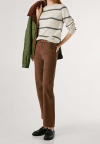 Femme portant un pull crème rayé, un pantalon marron, des mocassins noirs et tenant une veste matelassée verte sur son épaule.
