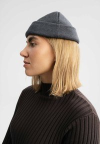 Grijze gebreide beanie met een ronde bovenkant en geribbelde textuur, gedragen door een persoon met steil blond haar, gecombineerd met een zwarte geribbelde trui.