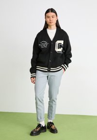 Cardigan negru cu dungi albe și logo, asociat cu pantaloni gri deschis. Pantofi negri cu un model maro. Fundal uniform.