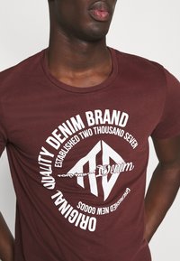 Camiseta de algodón burdeos con un diseño gráfico circular blanco que incluye texto, como "Marca de Denim de Calidad" y un logo emblemático.