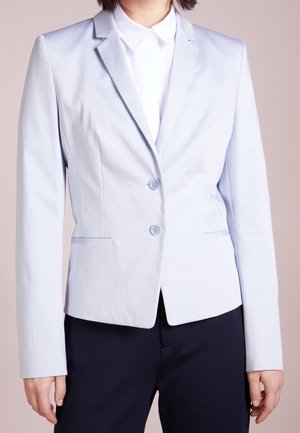 Femme portant un blazer ajusté bleu clair avec deux boutons sur une chemise blanche à col et un pantalon foncé, debout devant un fond uni.