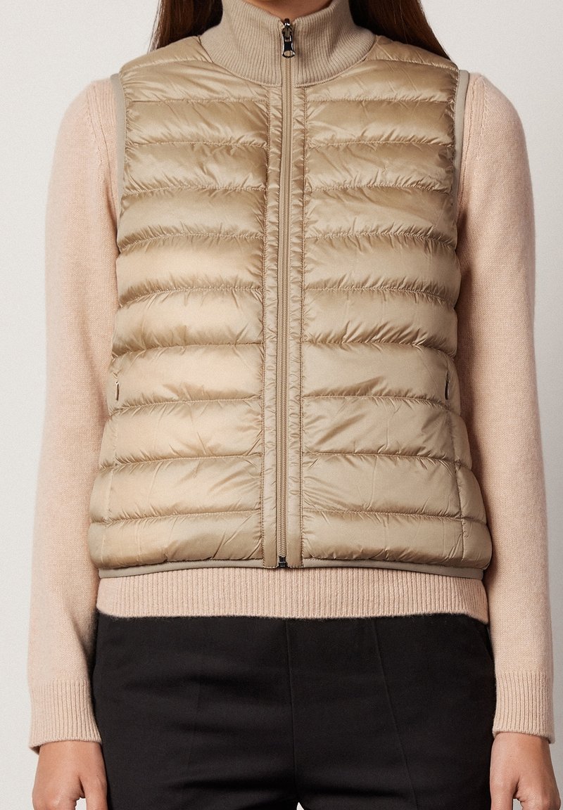 Nainen, joka pukeutuu vaaleanbeigeen tikattuun vetoketjulliseen liiviin vaaleanpunaisen pitkähihasen neuleen ja mustien housujen päällä, seisoo yksinkertaista taustaa vasten.