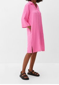 Rosa Leinenkleid mit Kragen, kurzen Ärmeln, seitlichen Taschen und seitlichen Schlitzen. Getragen mit schwarzen Riemchensandalen und einem strukturierten Stoff.