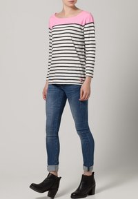 Striped long-sleeve shirt featuring a pink neckline, gray and white stripes, and a small logo detail at the hem, worn with blue jeans and black boots.