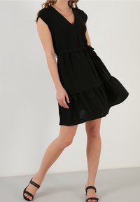 Svart kort klänning med V-ringning, korta ärmar och lagerad fåll. Har ett tygbälte för en figursydd siluett och en strukturerad yta.