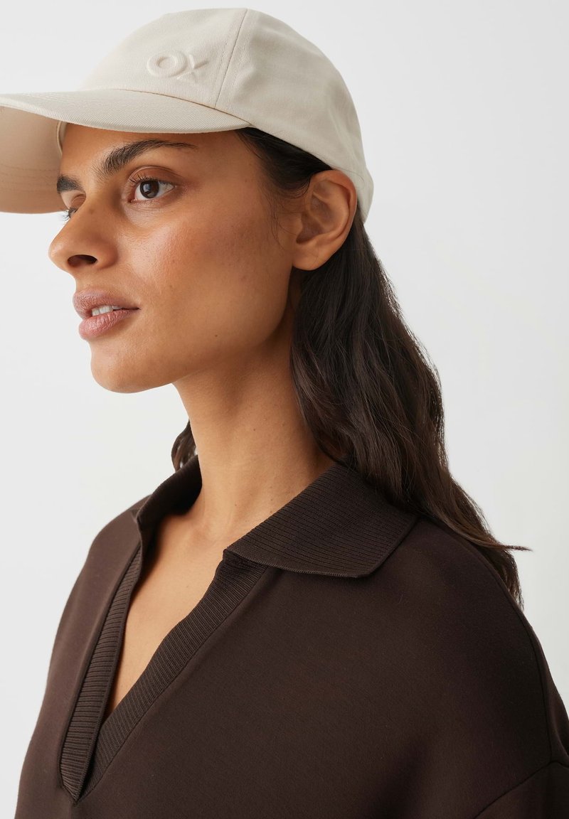 Frau mit dunklem Haar, die eine beige Kappe und ein dunkelbraunes Hemd mit Kragen trägt, blickt leicht nach oben vor einem einfachen hellen Hintergrund.