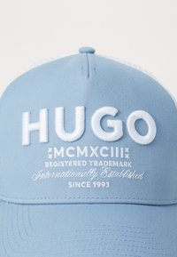 Gaiši zils beisbola cepure ar paaugstinātu balto izšūto "HUGO" logo, preču zīmju detaļām un izveides datumu 1993. Gluda auduma tekstūra.