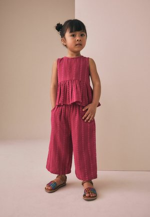 Noor tumeda peaga tüdruk kuklas, kandes varrukateta roosat mustrilist topi ja laiade säärtega pükse, seistes käed taskutes.