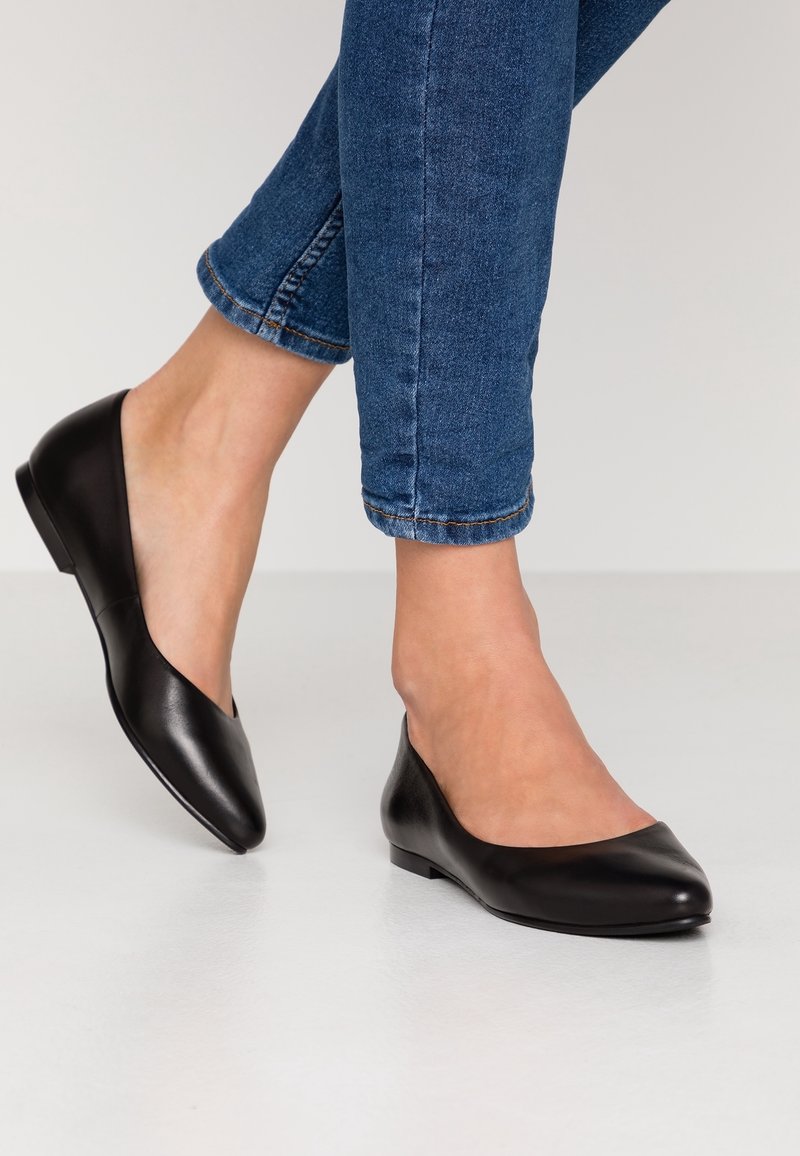 Jambes portant un jean bleu et des ballerines en cuir noir à bouts pointus, debout sur un sol clair.