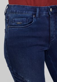 Vaqueros de mezclilla azul oscuro con un bolsillo frontal, cierre de botón metálico y detalles sutiles de costura en los laterales. Textura suave.