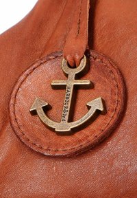 Étiquette en cuir marron avec un design circulaire, comportant un emblème d'ancre en bronze et un texte gravé "HARBOUR & 2nd." Texture lisse et bords cousus.