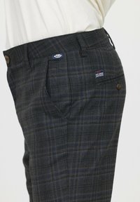 Pantalons à carreaux gris avec des accents bleus et jaunes, présentant des poches avant, une taille avec un bouton, et une étiquette sur le côté avant.