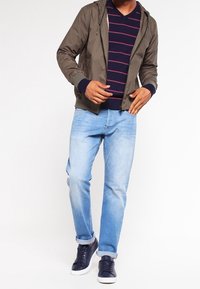 Veste bomber à capuche verte portée sur un pull à rayures bleu marine, associée à un jean bleu clair et des baskets bleu marine. Design décontracté et contemporain.