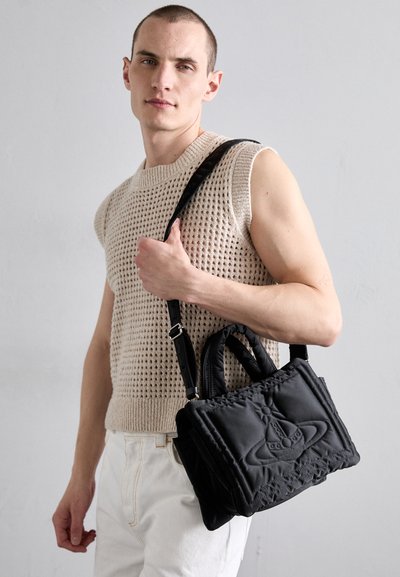 Bolso de mano negro acolchado con asas dobles y una correa desmontable. Presenta un diseño bordado y una textura suave. Lleva puesto una modelo con un top de punto.
