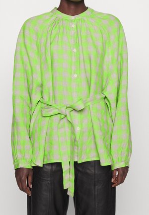 Personne portant un chemisier à manches longues à carreaux vert néon et beige avec des boutons sur le devant et une ceinture nouée à la taille, associé à un pantalon noir.