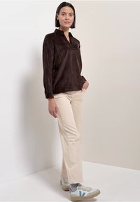 Donna in piedi con le mani nelle tasche, vestita con un top a maniche lunghe marrone scuro, pantaloni beige e sneakers bianche, su uno sfondo semplice.