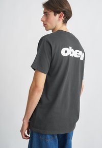 T-shirt de algodão cinza com um logótipo "obey" branco nas costas. Mangas curtas, gola redonda, corte relaxado. Usado com calções de ganga azuis.