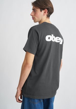 Szary bawełnowy t-shirt z białym logo 'obey' na plecach. Krótkie rękawy, okrągły dekolt, luźny krój. Noszony z niebieskimi dżinsowymi szortami.
