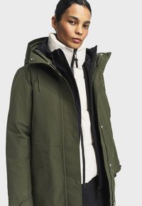 Parka vert olive avec un zip à l'avant, une capuche et de nombreuses poches. Superposé à une doudoune crème avec un col haut et des accents noirs.