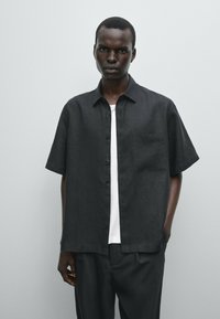 Camicia a maniche corte, con bottoni, in lino nero, dotata di taschino sul petto e vestibilità rilassata, indossata sopra una t-shirt bianca. Texture liscia.