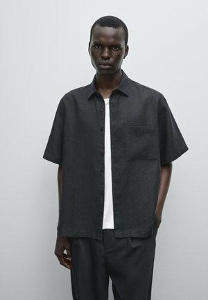 Short-sleeve, button-up shirt in black linen with a chest pocket and a relaxed fit, layered over a white t-shirt. Smooth texture.