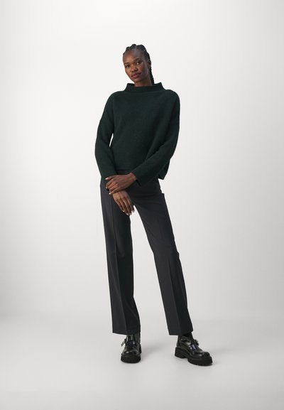 Maglione verde scuro, texturizzato, con collo alto, abbinato a pantaloni scuri e stivaletti neri con tacco a piattaforma. La modella sta in piedi con sicurezza, con le braccia incrociate.