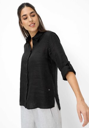 Donna con capelli lunghi e scuri che indossa una camicia nera con bottoni e maniche arrotolate e pantaloni grigi chiari su uno sfondo bianco.