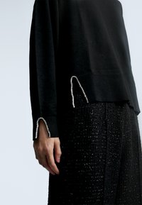 Černý pletený svetr s ribrovou texturou a stříbrnými ozdobami na rukávech, doplněný o texturované černé kalhoty.