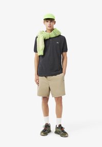 Grå pikétröja, beigefärgade shorts, grön keps och stickad grön scarf. Bär svarta och gröna sneakers med vita strumpor. Lacoste-loggan synlig.