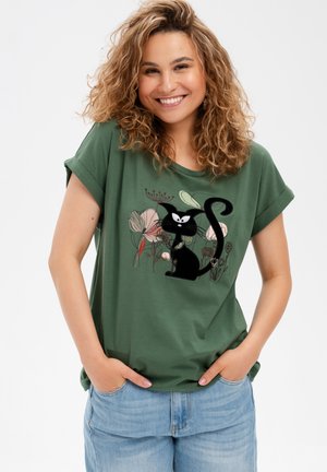 Uśmiechająca się kobieta z kręconymi włosami, ubrana w zielony t-shirt z czarnym kotem i kwiatowym wzorem, ręce w kieszeniach, jasnoniebieskie dżinsy, białe tło.