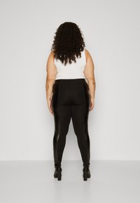 Black high-waisted leggings with faux leather side panels, paired with a white sleeveless top. Model stands with back to the camera.