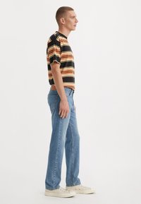 Gestreept T-shirt in zwart, oranje en geel, gecombineerd met lichtblauwe rechte jeans. Model draagt beige lage sneakers.