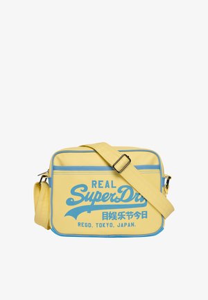 Żółta prostokątna torba na ramię z niebieskimi lamówkami i regulowanym paskiem, z niebieskim tekstem "Real SuperDry" i japońskimi znakami z przodu.