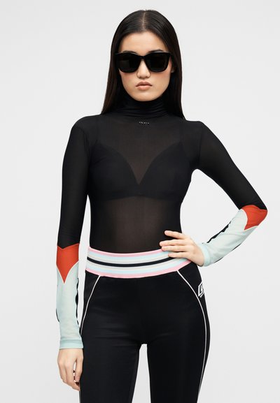 Γυναίκα που φοράει μαύρο διάφανο μπλουζάκι με μακριά μανίκια πολύχρωμα, μαύρο αθλητικό παντελόνι με ριγέ μέση και μαύρα γυαλιά ηλίου, με τα χέρια στη μέση.