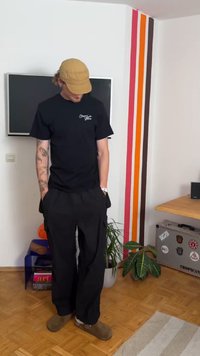Black graphic t-shirt, black loose-fitting pants, and brown clogs. Room features a TV, colorful wall stripes, and potted plants.