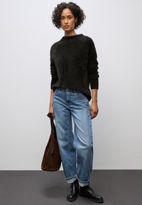 Schwarzer, strukturierter Pullover, locker sitzende blaue Jeans mit umgeschlagenen Säumen. Das Model hält eine braune Wildleder-Tasche und trägt schwarze Knöchelstiefel.
