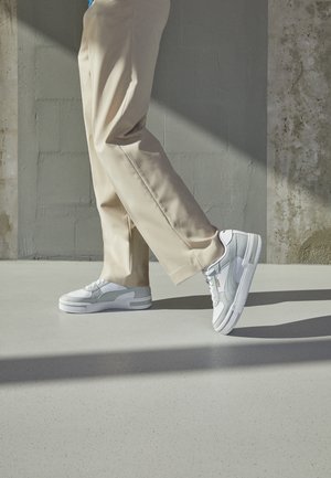 Personne portant un pantalon beige et des baskets blanches marchant sur une surface lisse et claire avec un mur en béton en arrière-plan.