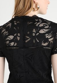 Černý krajkový top s květinovými vzory, krátkými rukávy, vroubkovaným výstřihem a otevřenými zády se zapínáním na zip. Texturovaný a jemný design.