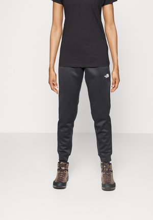 Osoba ubrana w czarną koszulkę z krótkim rękawem, ciemnoszare spodnie dresowe The North Face oraz brązowe buty turystyczne, stojąca na jednolitym jasnym tle.
