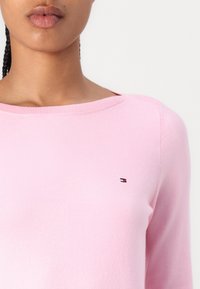 Lys pink, strikket genser med vid halslinning og lange ermer. Har en liten, brodert logo på venstre bryst. Myk tekstur.