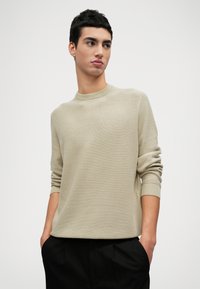 Helle beigefarbene Strickjacke mit Rundhalsausschnitt und gerippten Bündchen, die ein strukturiertes Muster aufweist. Getragen über schwarzen Hosen, Hände in den Taschen.