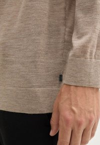 Beige Strickpullover mit weicher Textur und gerippten Bündchen. Verfügt über ein kleines dunkles Etikett am Saum zur Markenidentifikation.