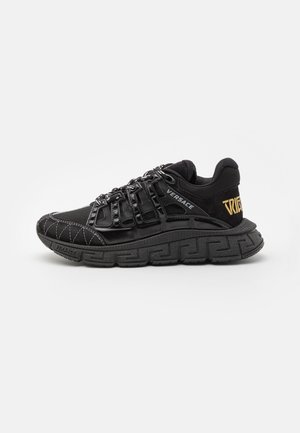 Sneaker atletico nero con tomaia textured, sistema di allacciatura prominente e suola in gomma con un motivo a chiave greca. Gli accenti includono il logo del marchio.