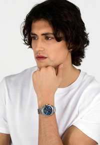 Orologio in metallo argento con quadrante blu, caratterizzato da tre sottoquadranti, una lunetta testurizzata e un bracciale a maglie in metallo, indossato su un polso contro una camicia bianca.