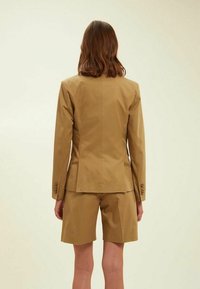 Blazer beige cintré avec une coupe droite, associé à un short assorti ; présente des accents de boutons sur les manches et une texture de tissu lisse.