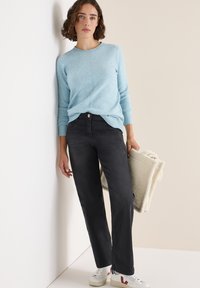 Hellblauer gestrickter Pullover, schwarze gerade Jeans und weiße Sneakers mit roten Akzenten. Hält eine cremefarbene strukturierte Decke.