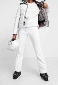 Vit isolerad jacka med grå detaljer, vit långärmad topp och vita skidbyxor. Svarta skidboots och en vit hjälm som hålls i handen.